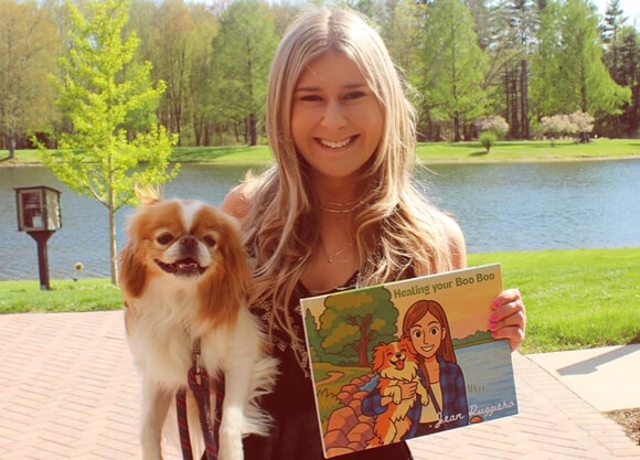 Student holds her dog in one hand, a book in the other, and smiles for a photo.
